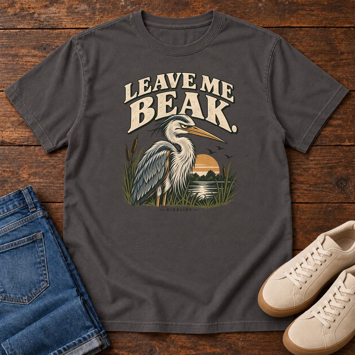 Leave Me Beak T-Shirt in Graphite, front laid flat on wood, featuring a gray, long-billed wading bird among reeds beside a sunset, beneath cream lettering.