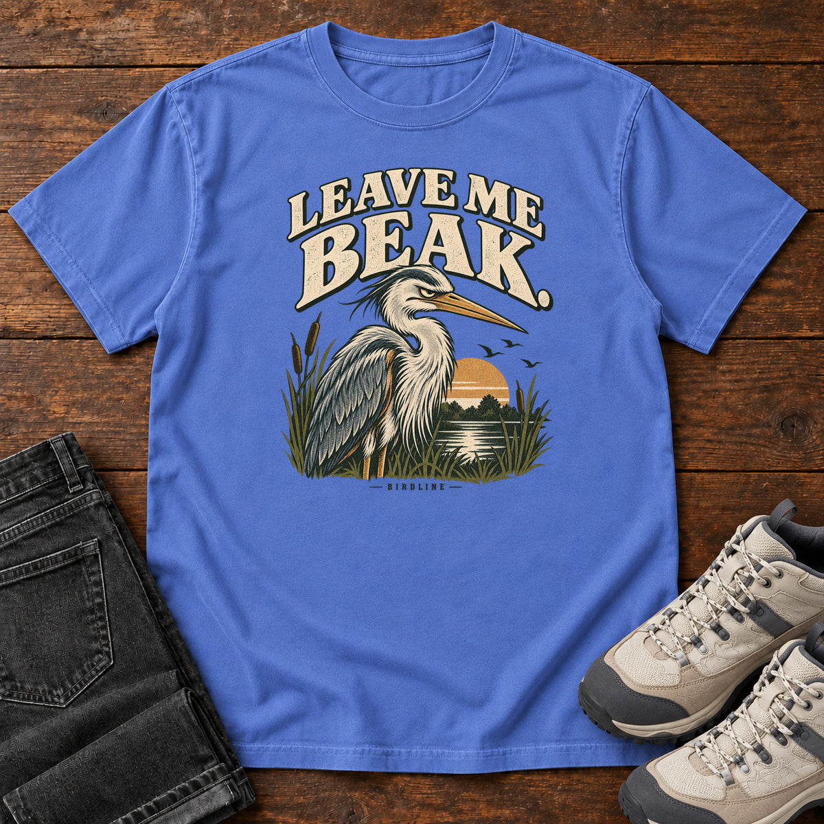 Leave Me Beak T-Shirt in Mystic Blue, front laid flat on wood, featuring a gray, long-billed wading bird among reeds beside a sunset, beneath cream lettering.