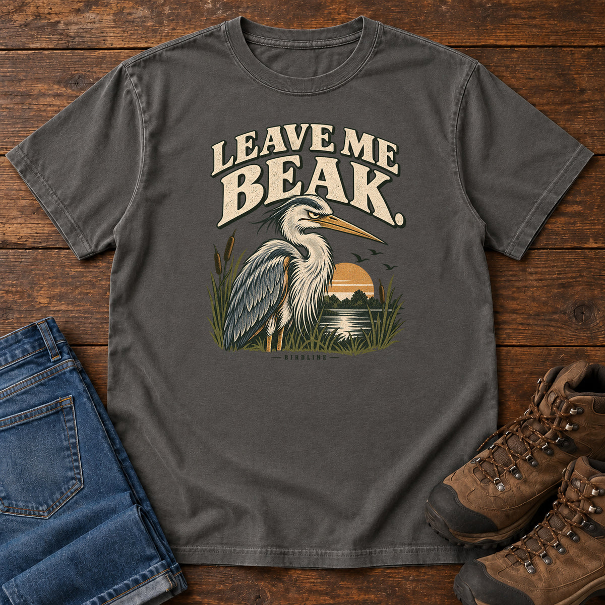 Leave Me Beak T-Shirt in Pepper, front laid flat on wood, featuring a gray, long-billed wading bird among reeds beside a sunset, beneath cream lettering.