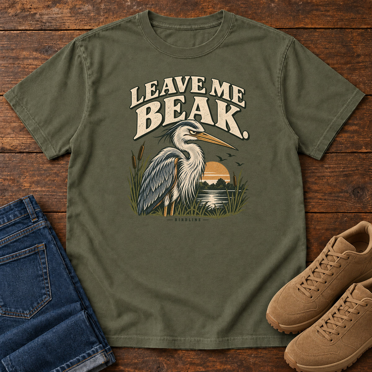 Leave Me Beak T-Shirt in Sage, front laid flat on wood, featuring a gray, long-billed wading bird among reeds beside a sunset, beneath cream lettering.