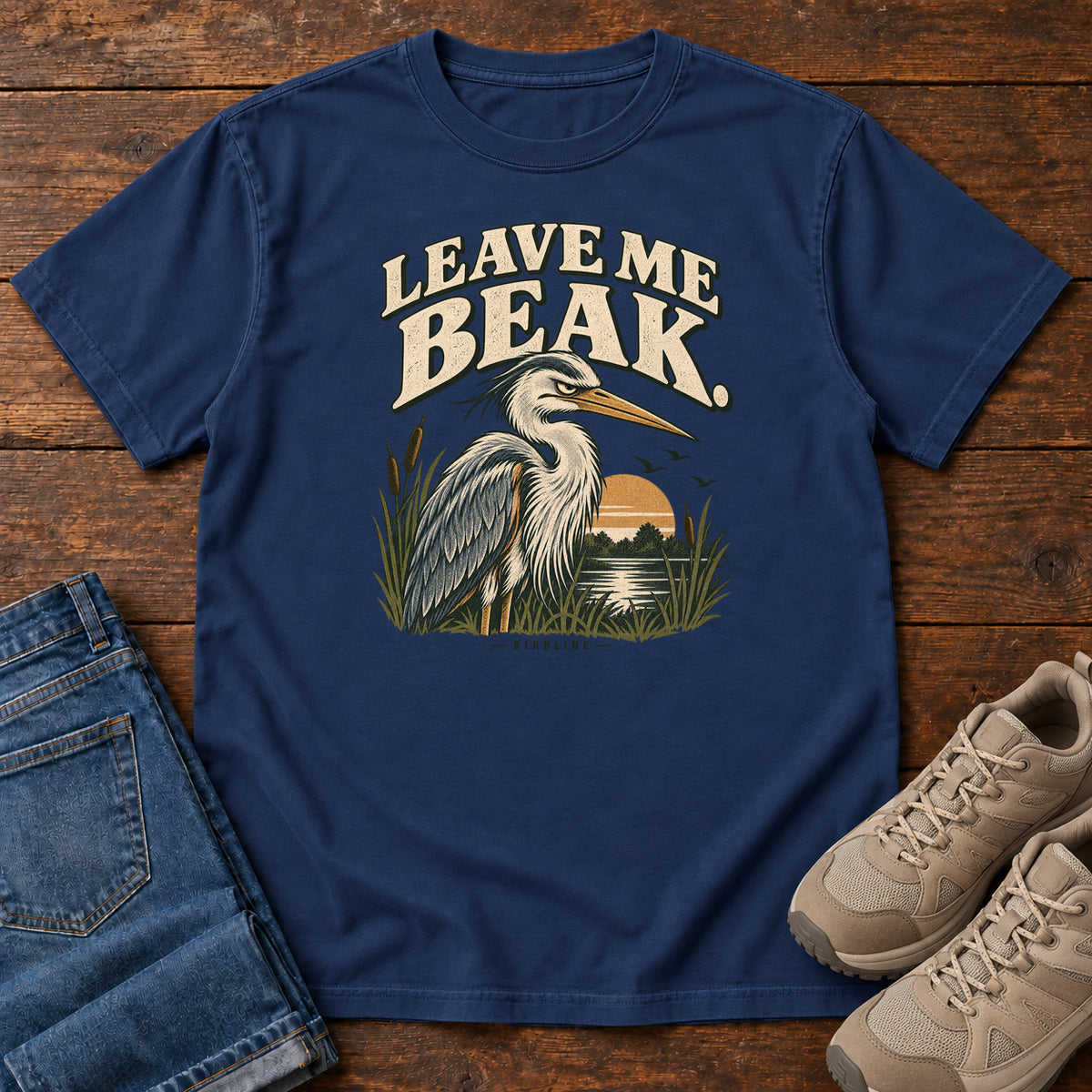 Leave Me Beak T-Shirt in True Navy, front laid flat on wood, featuring a gray, long-billed wading bird among reeds beside a sunset, beneath cream lettering.