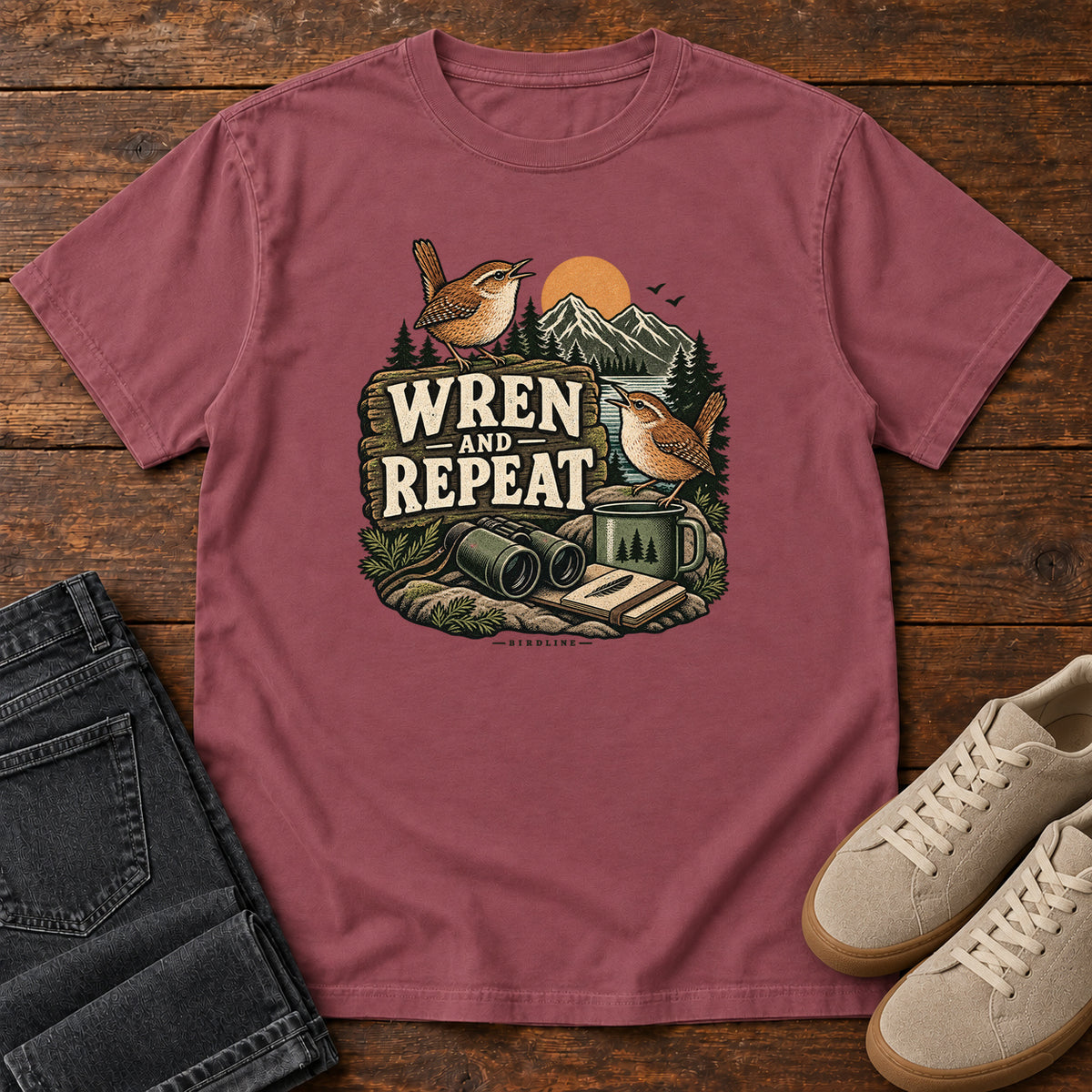 Brick Wren and Repeat T-Shirt with two small brown birds, binoculars and a camp mug around Wren and Repeat lettering, front flat lay on wood.