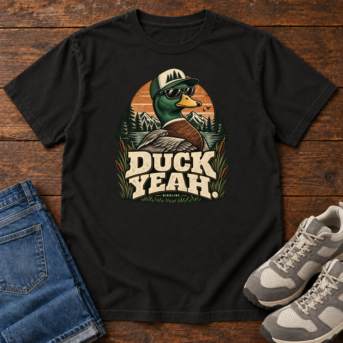 Black Duck Yeah T-Shirt with a green-headed duck wearing a cap and sunglasses above bold cream lettering, front flat lay on wood.