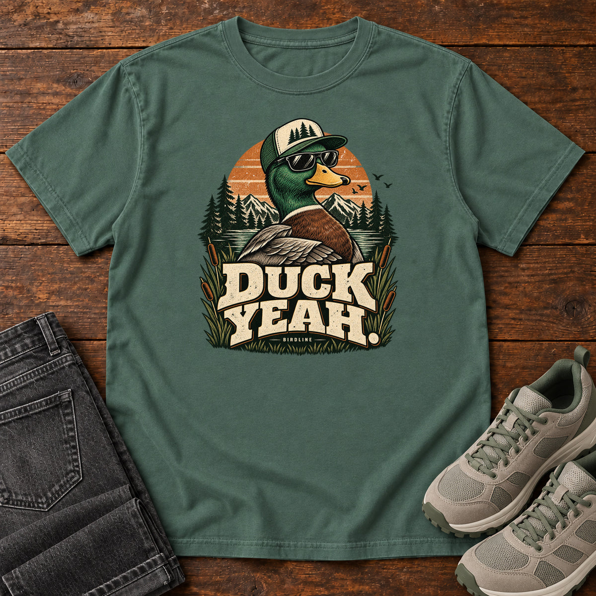 Blue Spruce Duck Yeah T-Shirt with a green-headed duck wearing a cap and sunglasses above bold cream lettering, front flat lay on wood.
