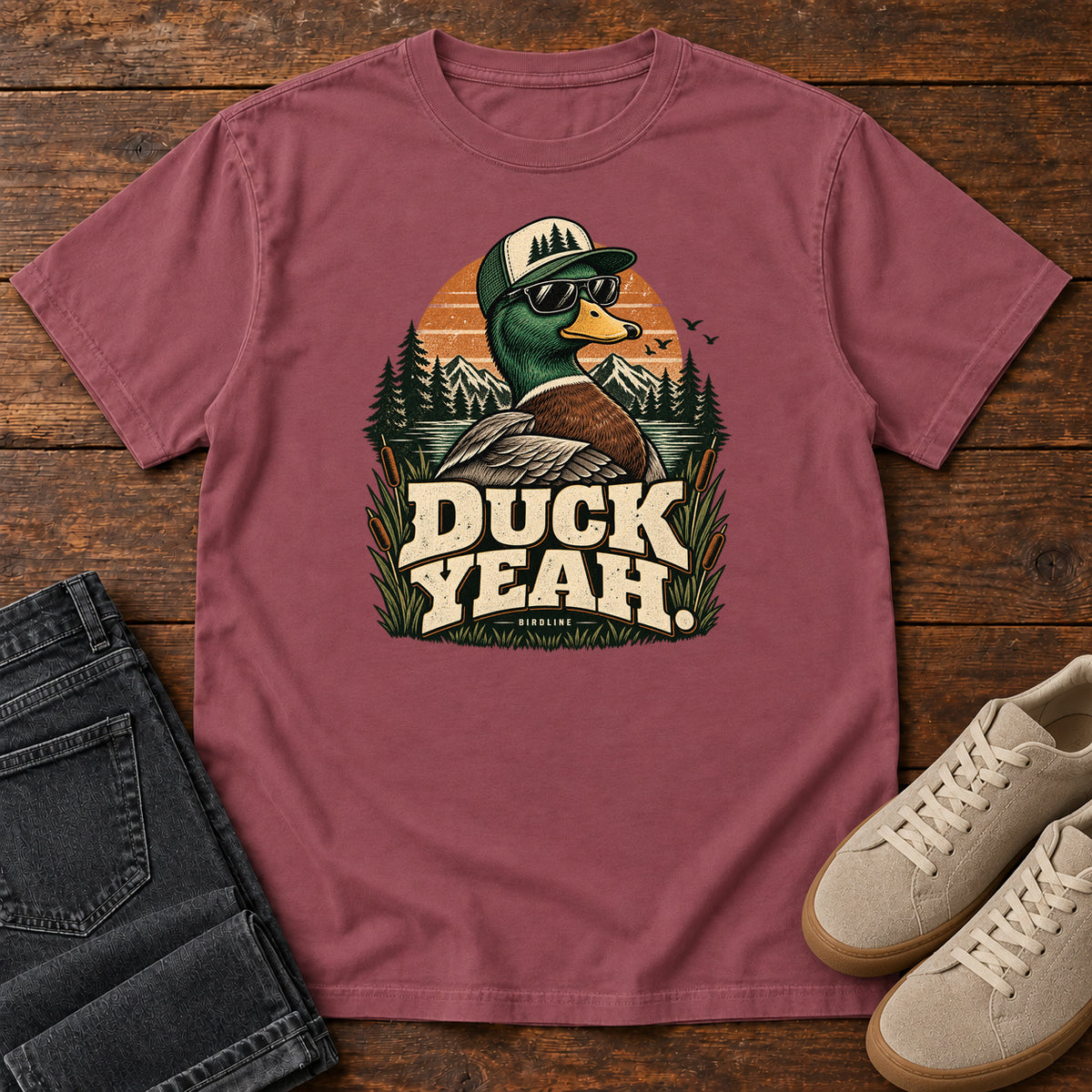 Brick Duck Yeah T-Shirt with a green-headed duck wearing a cap and sunglasses above bold cream lettering, front flat lay on wood.