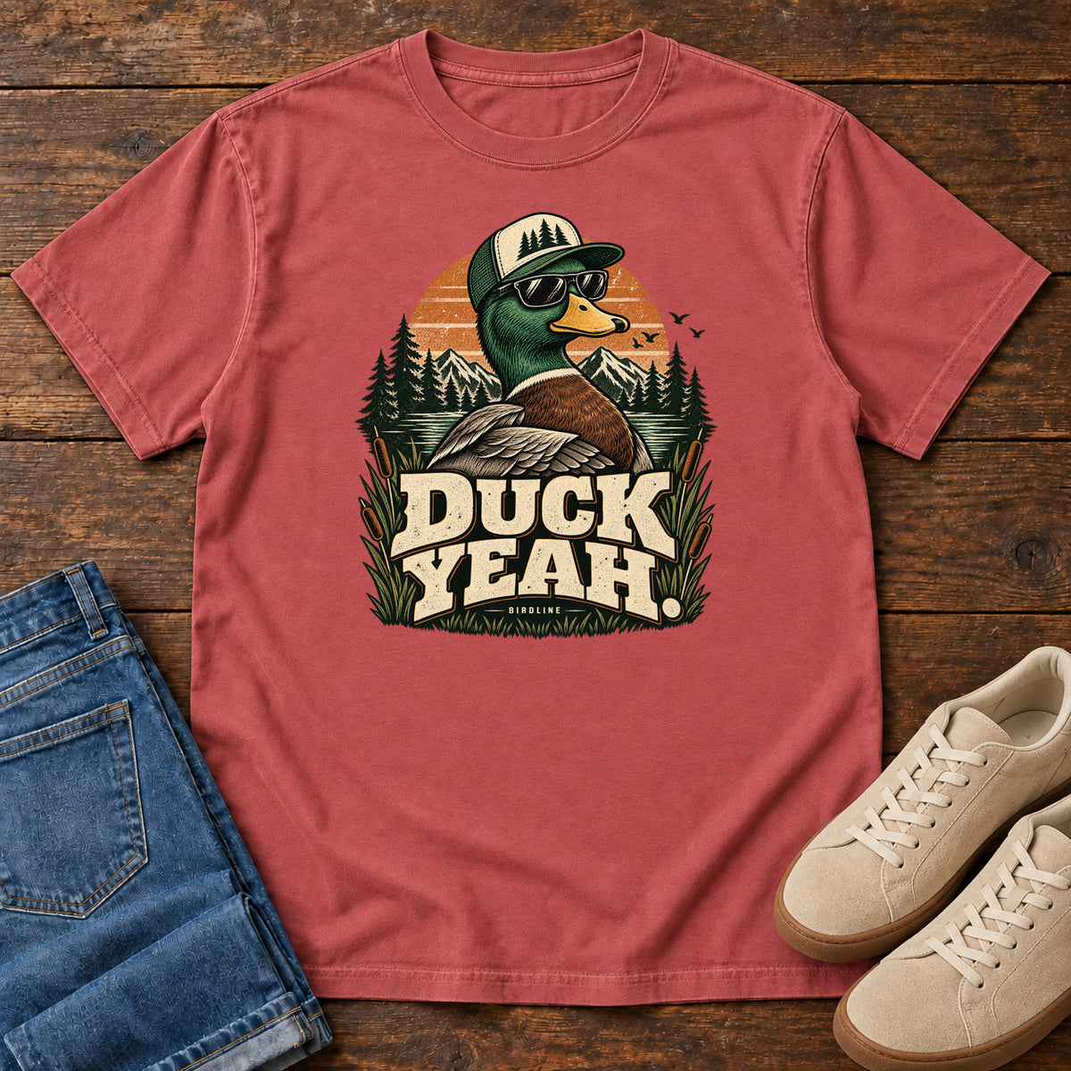 Crimson Duck Yeah T-Shirt with a green-headed duck wearing a cap and sunglasses above bold cream lettering, front flat lay on wood.