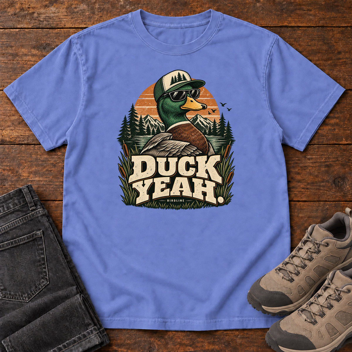 Flo Blue Duck Yeah T-Shirt with a green-headed duck wearing a cap and sunglasses above bold cream lettering, front flat lay on wood.
