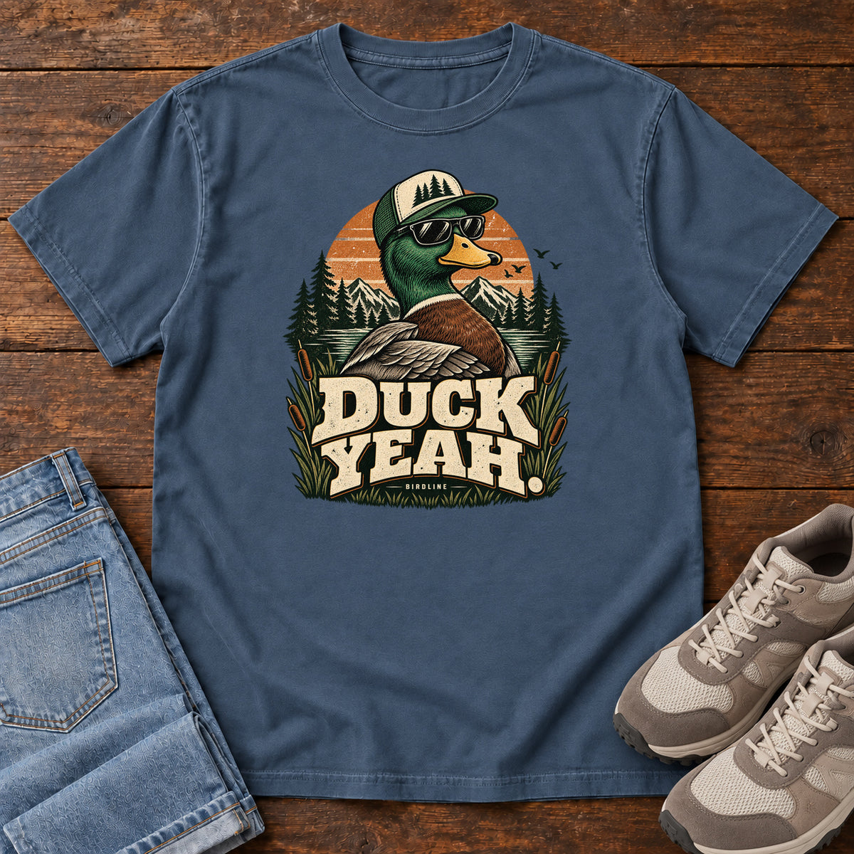 Midnight Duck Yeah T-Shirt with a green-headed duck wearing a cap and sunglasses above bold cream lettering, front flat lay on wood.