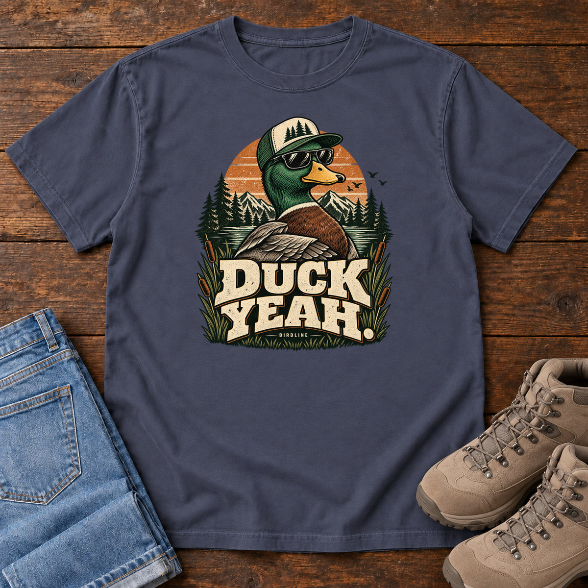 Navy Duck Yeah T-Shirt with a green-headed duck wearing a cap and sunglasses above bold cream lettering, front flat lay on wood.