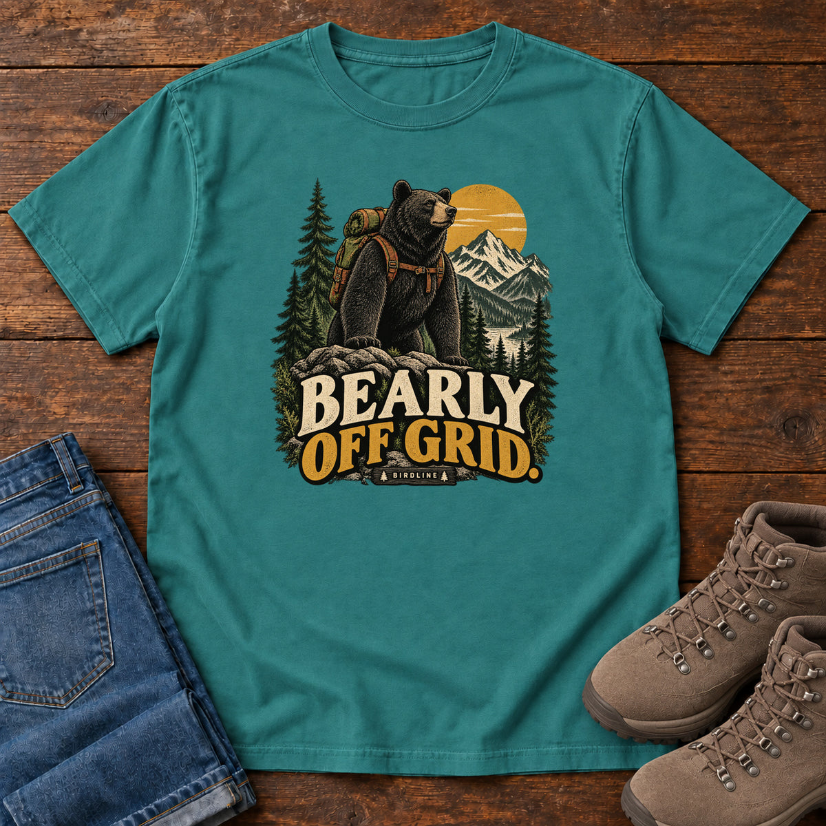 Emerald Bearly Off Grid T-Shirt with a black bear wearing a backpack before mountains, pines and a golden sun; front flat lay on wood.