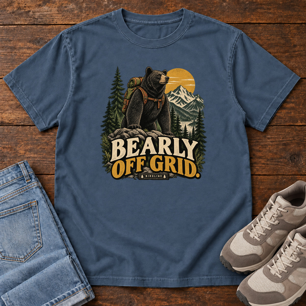 Midnight Bearly Off Grid T-Shirt with a black bear wearing a backpack before mountains, pines and a golden sun; front flat lay on wood.