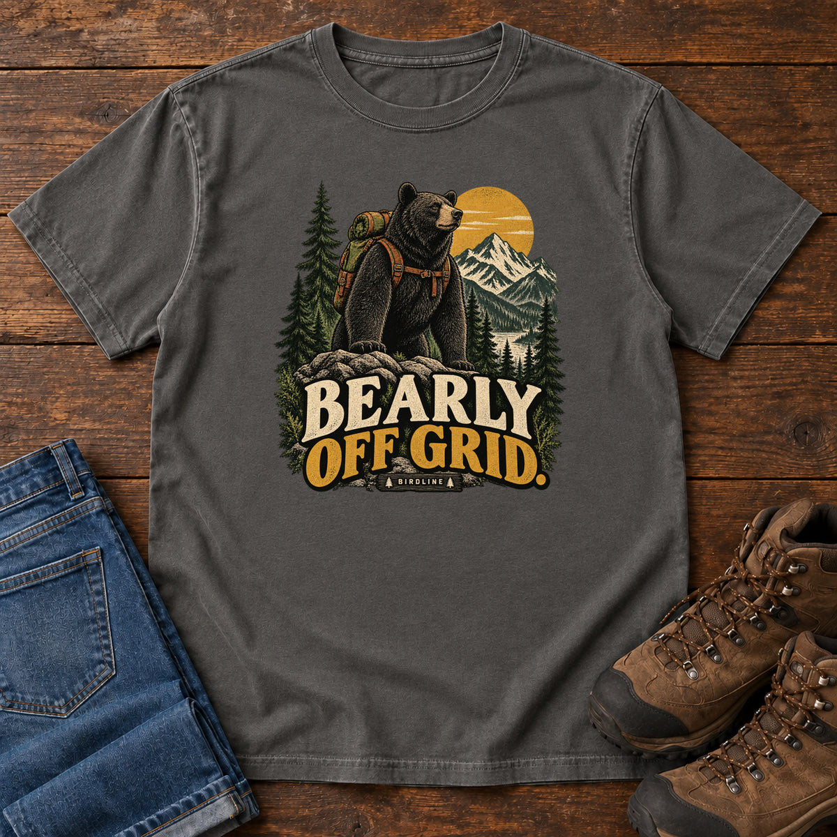 Pepper Bearly Off Grid T-Shirt with a black bear wearing a backpack before mountains, pines and a golden sun; front flat lay on wood.