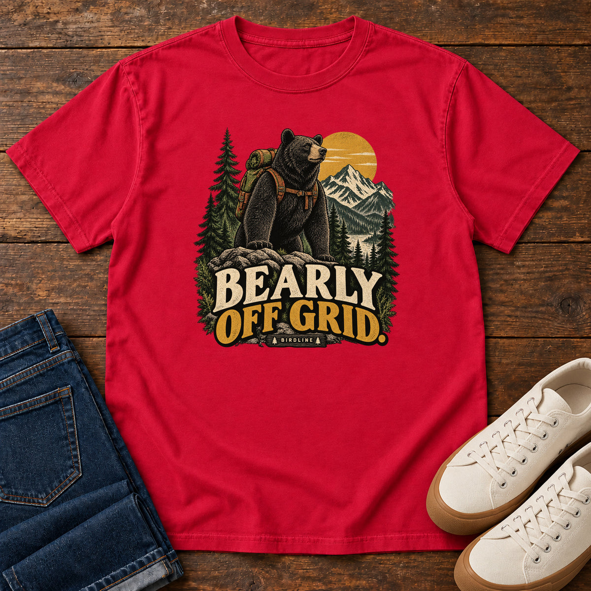 Red Bearly Off Grid T-Shirt with a black bear wearing a backpack before mountains, pines and a golden sun; front flat lay on wood.