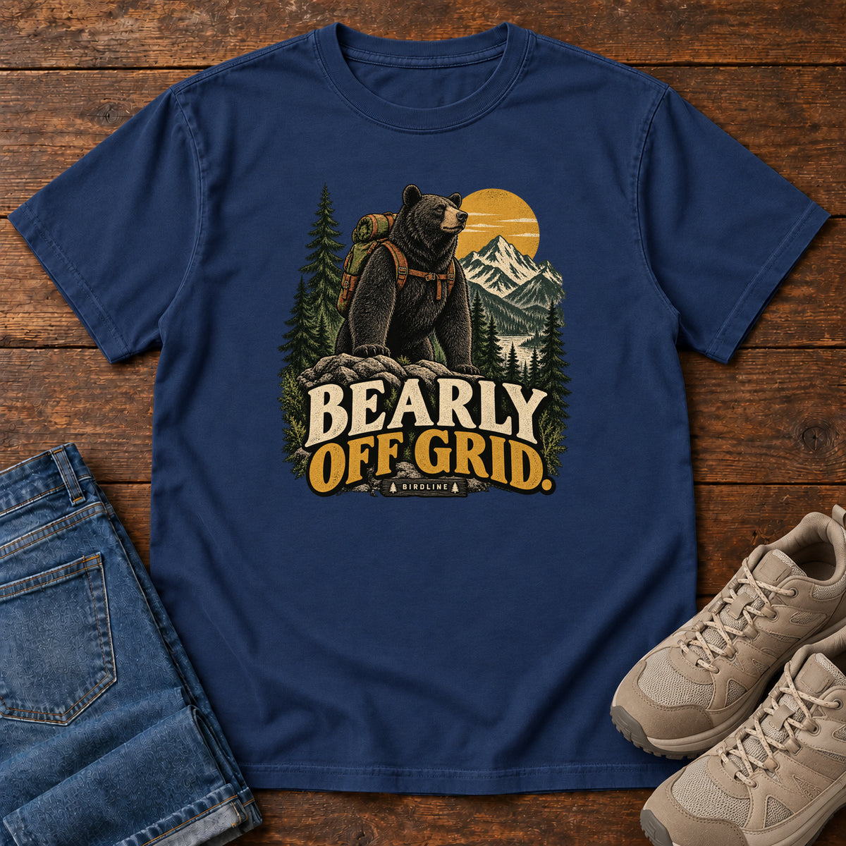 True Navy Bearly Off Grid T-Shirt with a black bear wearing a backpack before mountains, pines and a golden sun; front flat lay on wood.