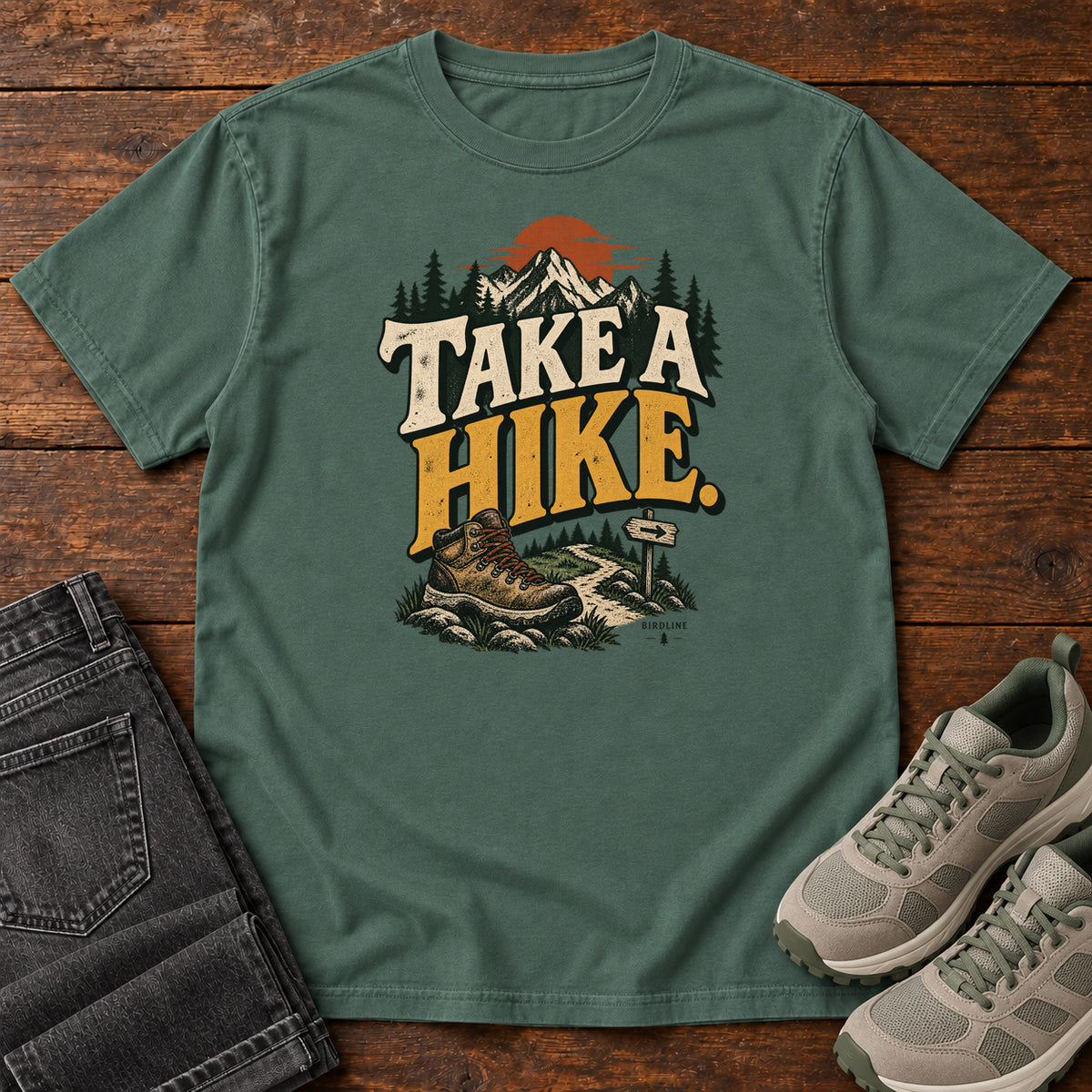 Blue Spruce Take a Hike T-Shirt with a hiking boot, mountain trail and orange sun around bold cream and gold lettering; front flat lay on wood.