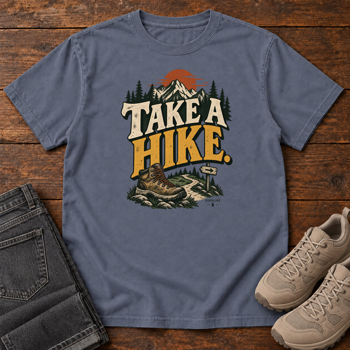 Denim Take a Hike T-Shirt with a hiking boot, mountain trail and orange sun around bold cream and gold lettering; front flat lay on wood.