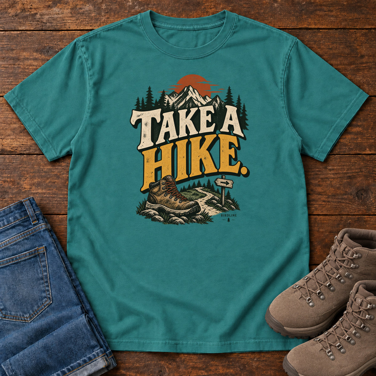Emerald Take a Hike T-Shirt with a hiking boot, mountain trail and orange sun around bold cream and gold lettering; front flat lay on wood.