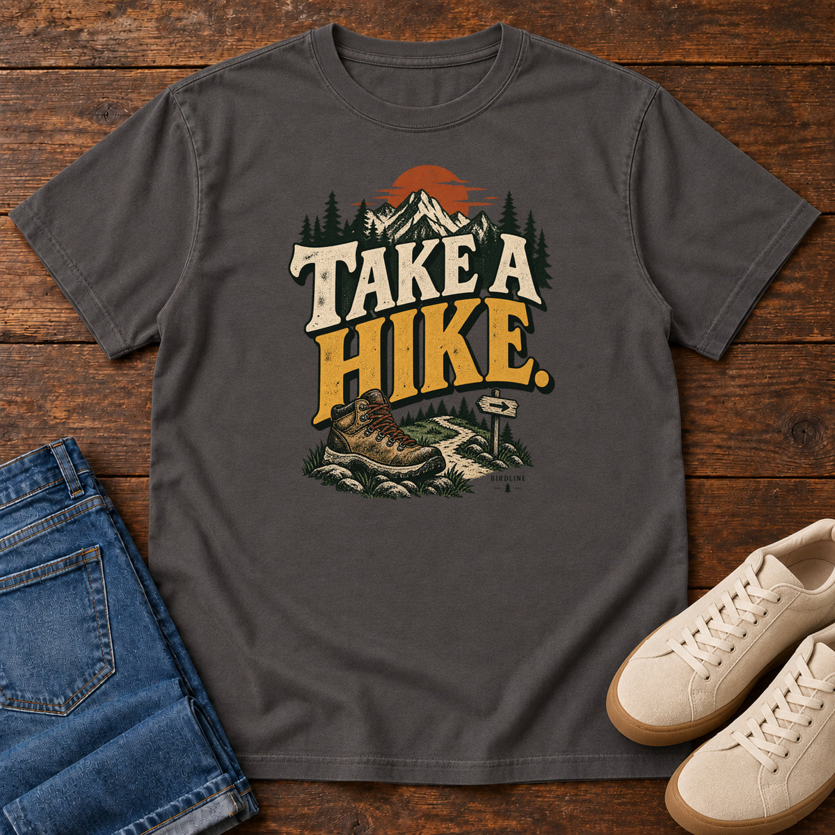 Graphite Take a Hike T-Shirt with a hiking boot, mountain trail and orange sun around bold cream and gold lettering; front flat lay on wood.
