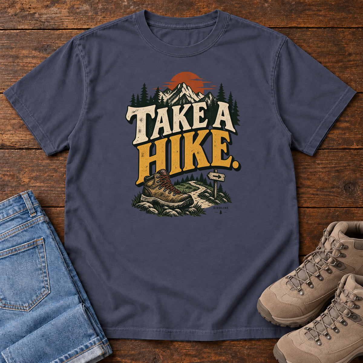 Navy Take a Hike T-Shirt with a hiking boot, mountain trail and orange sun around bold cream and gold lettering; front flat lay on wood.