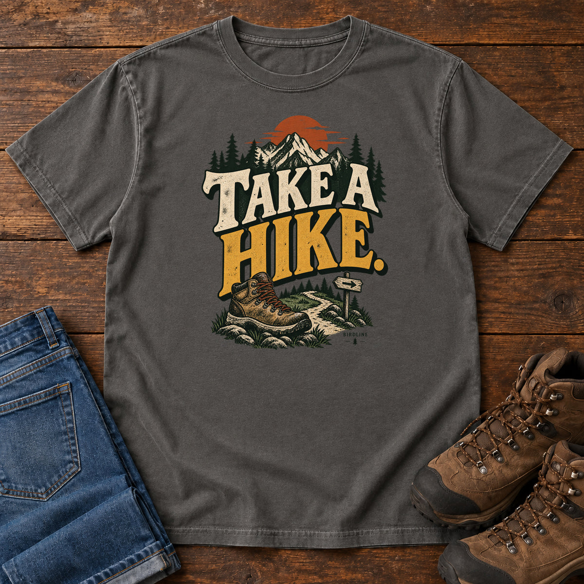 Pepper Take a Hike T-Shirt with a hiking boot, mountain trail and orange sun around bold cream and gold lettering; front flat lay on wood.