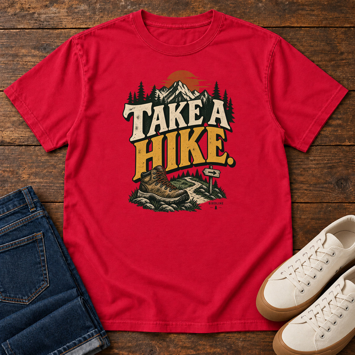 Red Take a Hike T-Shirt with a hiking boot, mountain trail and orange sun around bold cream and gold lettering; front flat lay on wood.