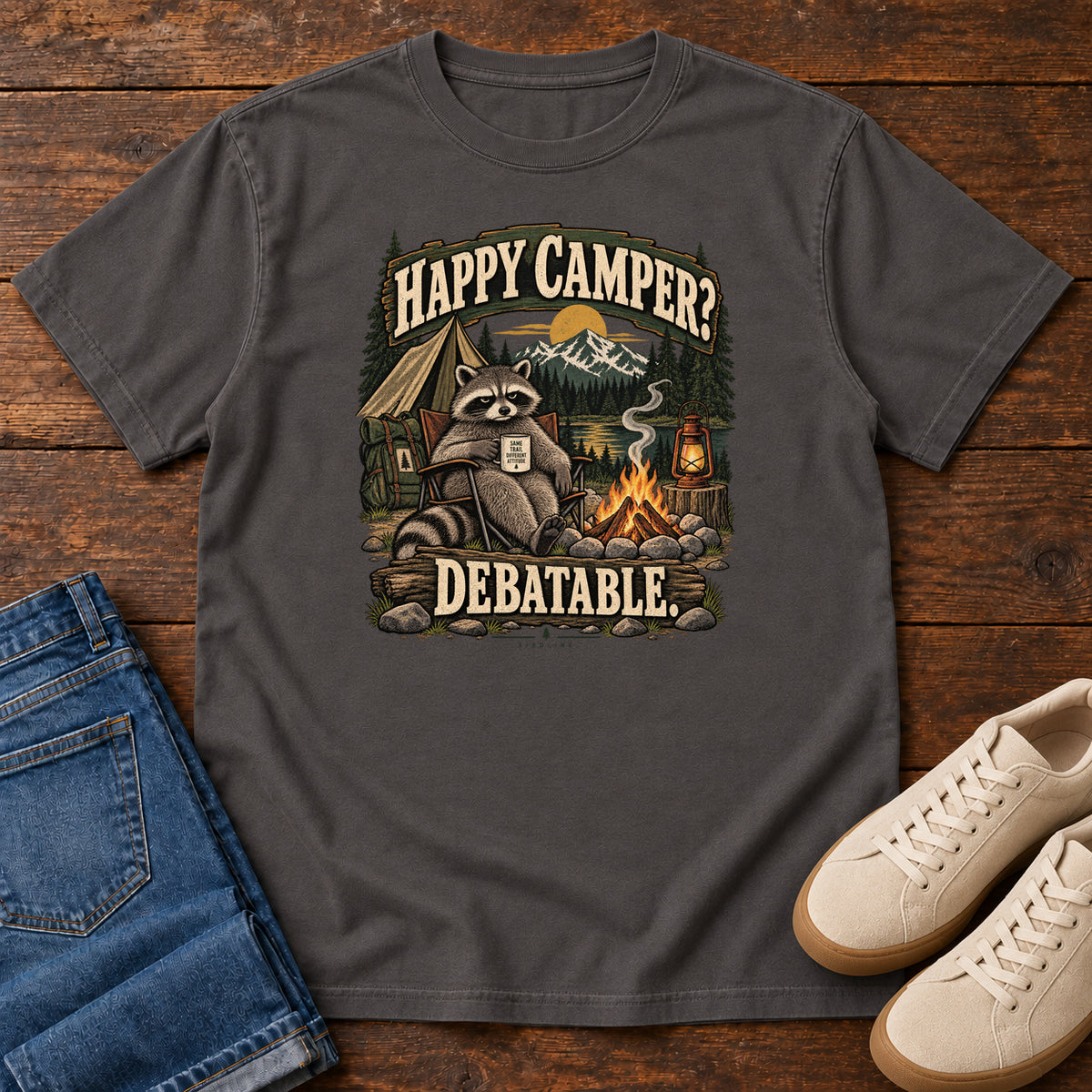 Graphite Happy Camper Debatable T-Shirt with a raccoon holding a mug beside a campfire, tent and lantern, front flat lay on wood.