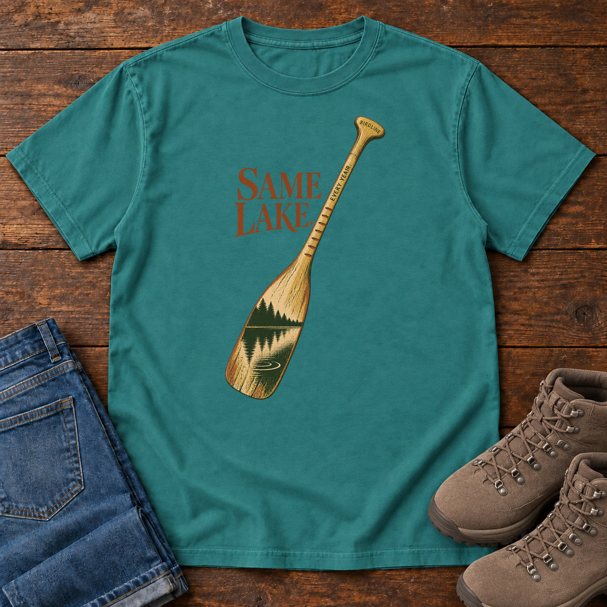 Emerald Same Lake Every Year T-shirt with a paddle and lake illustration, laid flat on a wooden surface.