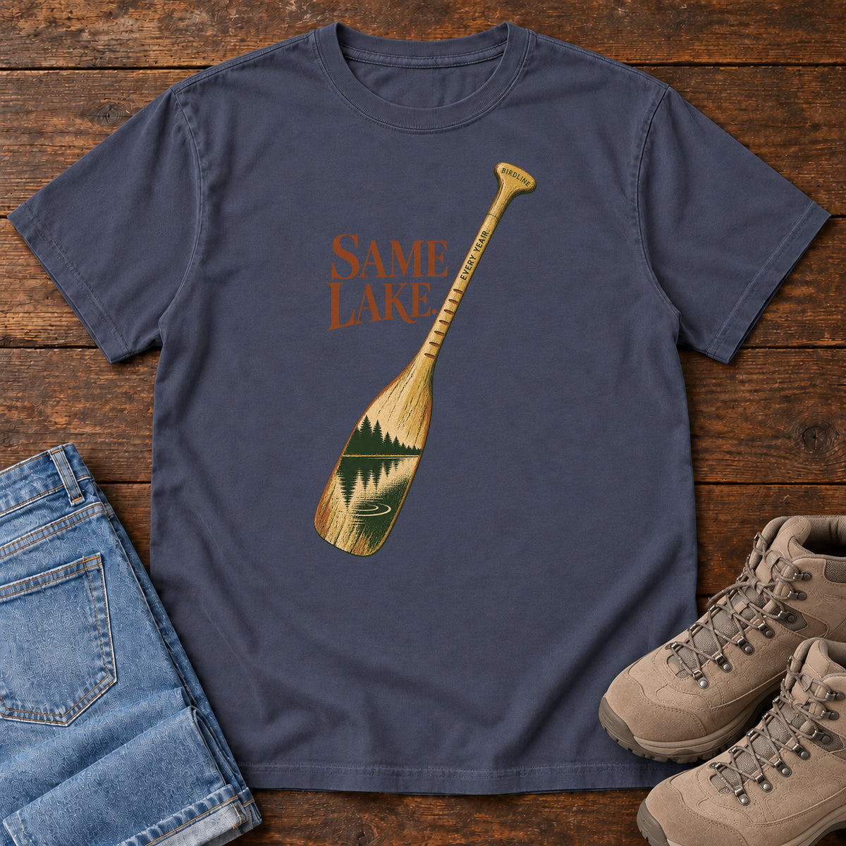 Navy Same Lake Every Year T-shirt with a paddle and lake illustration, laid flat on a wooden surface.