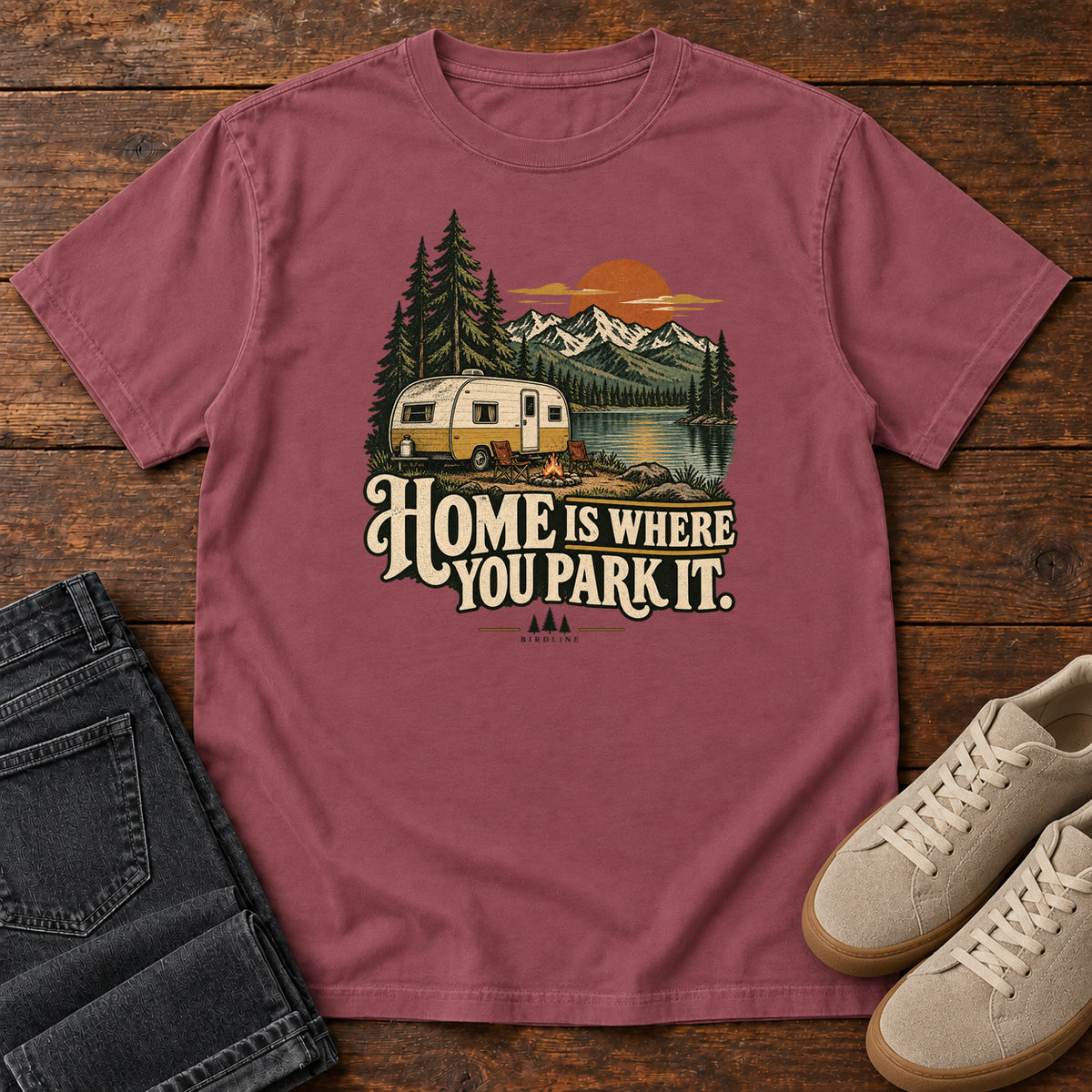 Home Is Where You Park It T-Shirt in Brick, front laid flat on wood, featuring a cream camper beside a mountain lake, pine trees and an orange sun above slogan lettering.