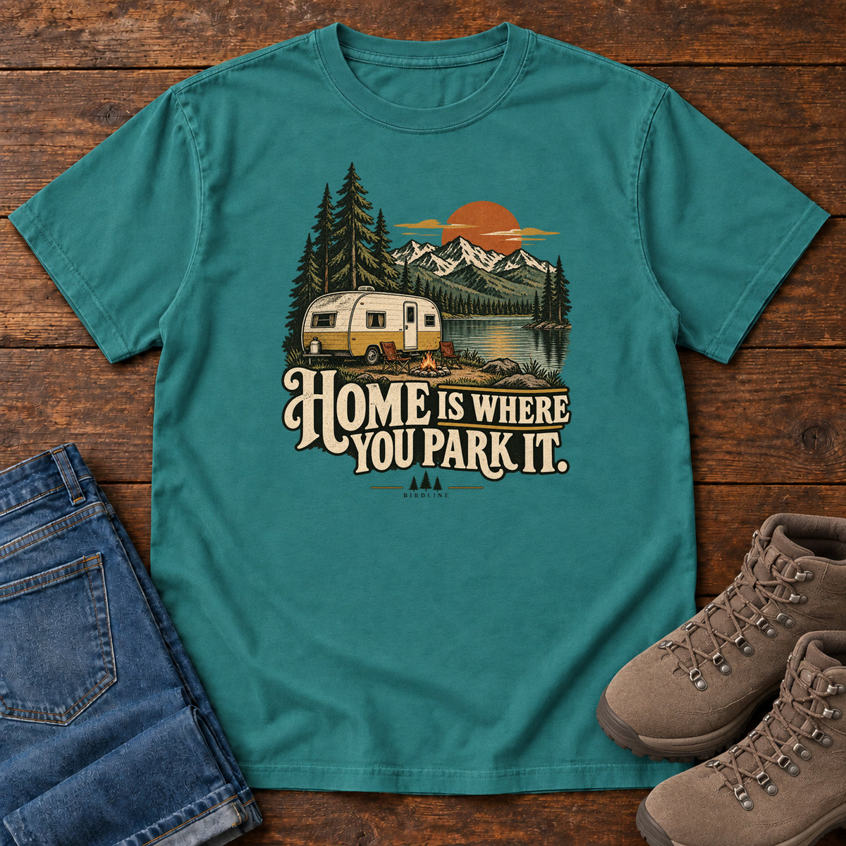 Home Is Where You Park It T-Shirt in Emerald, front laid flat on wood, featuring a cream camper beside a mountain lake, pine trees and an orange sun above slogan lettering.