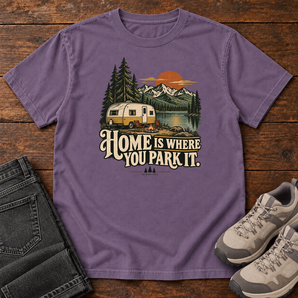 Home Is Where You Park It T-Shirt in Grape, front laid flat on wood, featuring a cream camper beside a mountain lake, pine trees and an orange sun above slogan lettering.