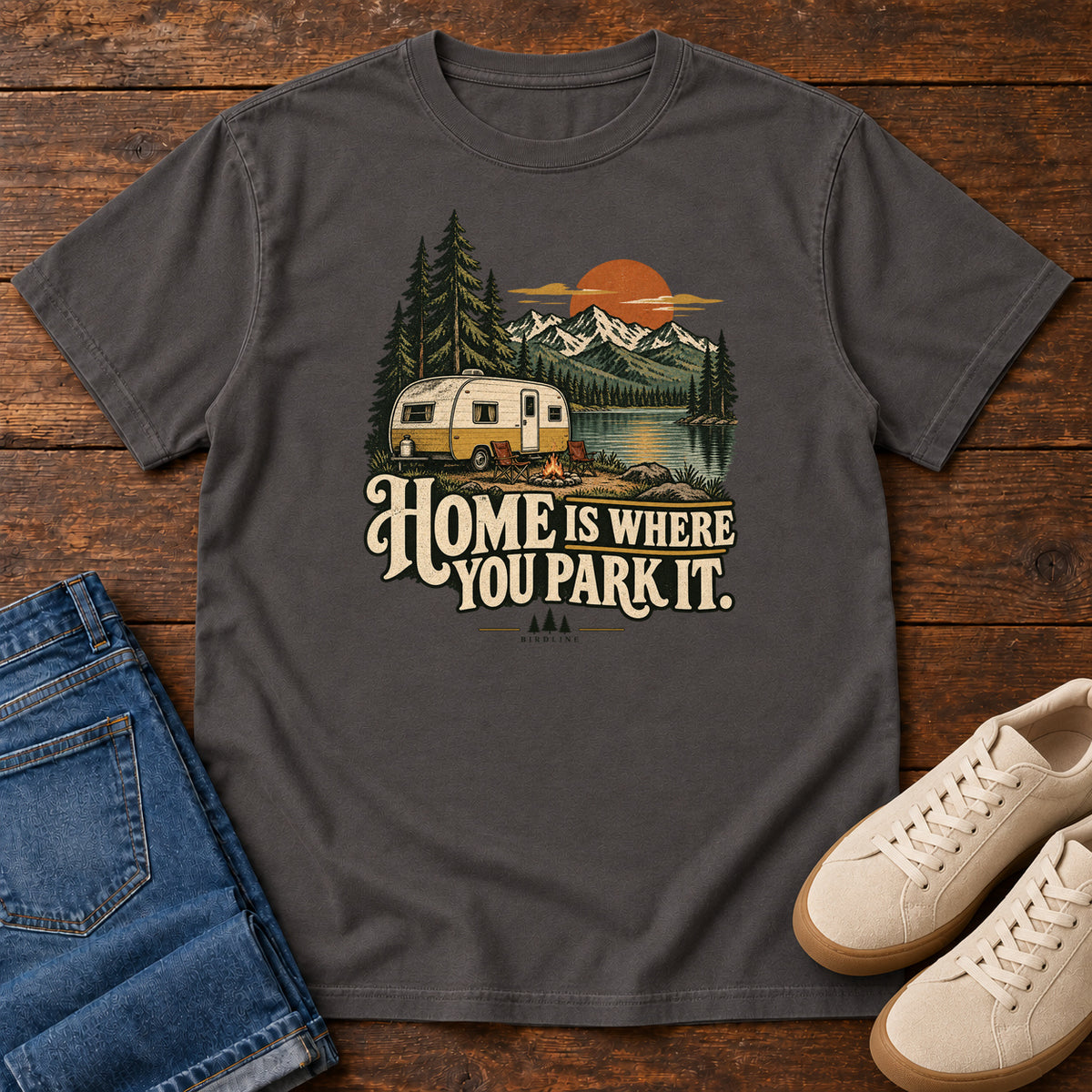 Home Is Where You Park It T-Shirt in Graphite, front laid flat on wood, featuring a cream camper beside a mountain lake, pine trees and an orange sun above slogan lettering.