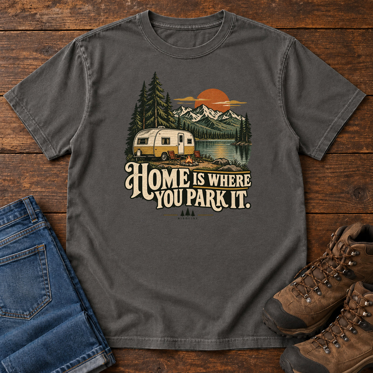 Home Is Where You Park It T-Shirt in Pepper, front laid flat on wood, featuring a cream camper beside a mountain lake, pine trees and an orange sun above slogan lettering.