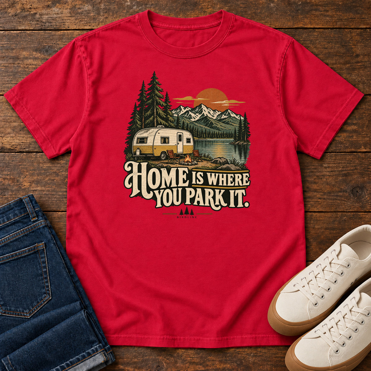 Home Is Where You Park It T-Shirt in Red, front laid flat on wood, featuring a cream camper beside a mountain lake, pine trees and an orange sun above slogan lettering.