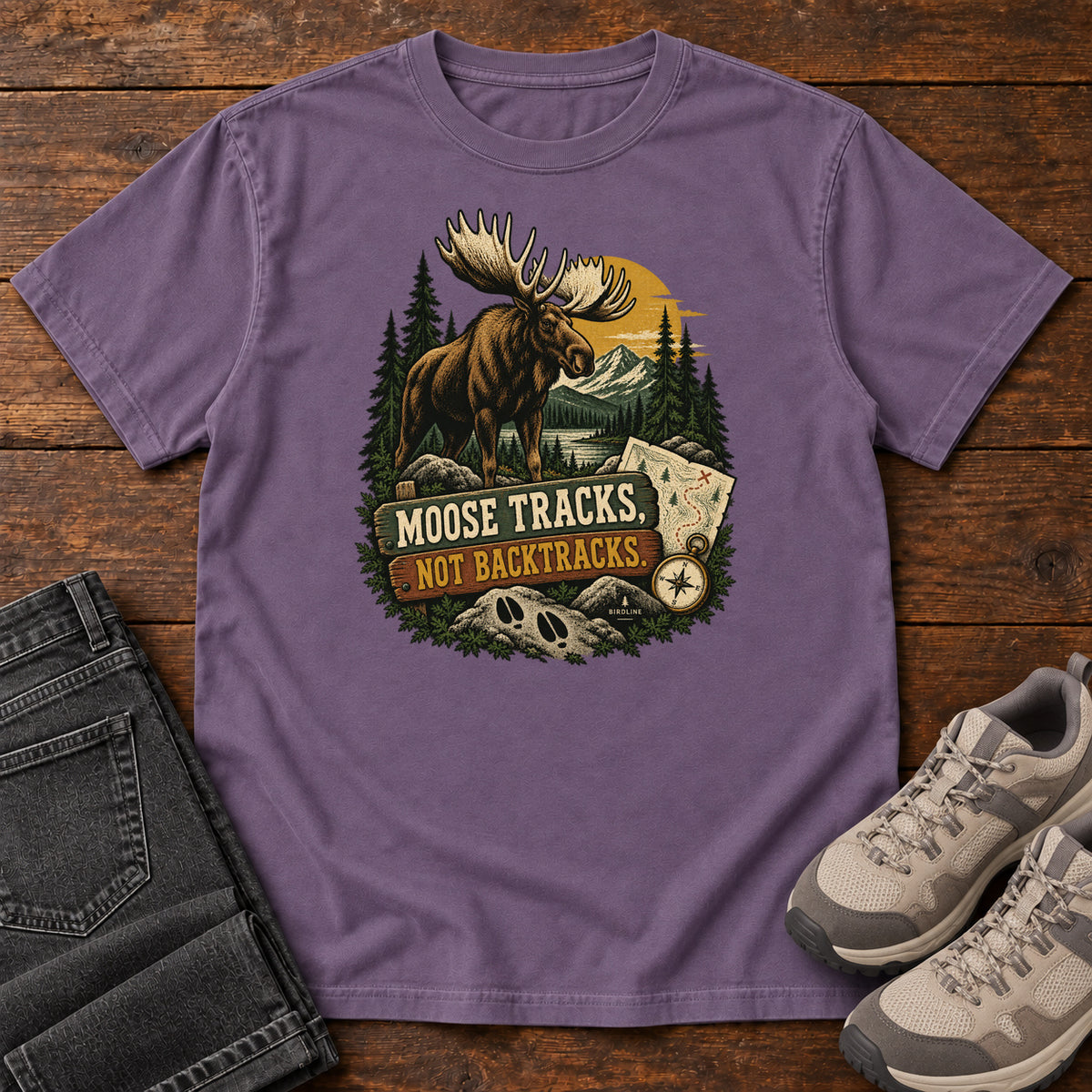 Grape Moose Tracks Not Backtracks T-Shirt with a moose in a mountain forest above a map, compass and cream and gold lettering, front flat lay on wood.