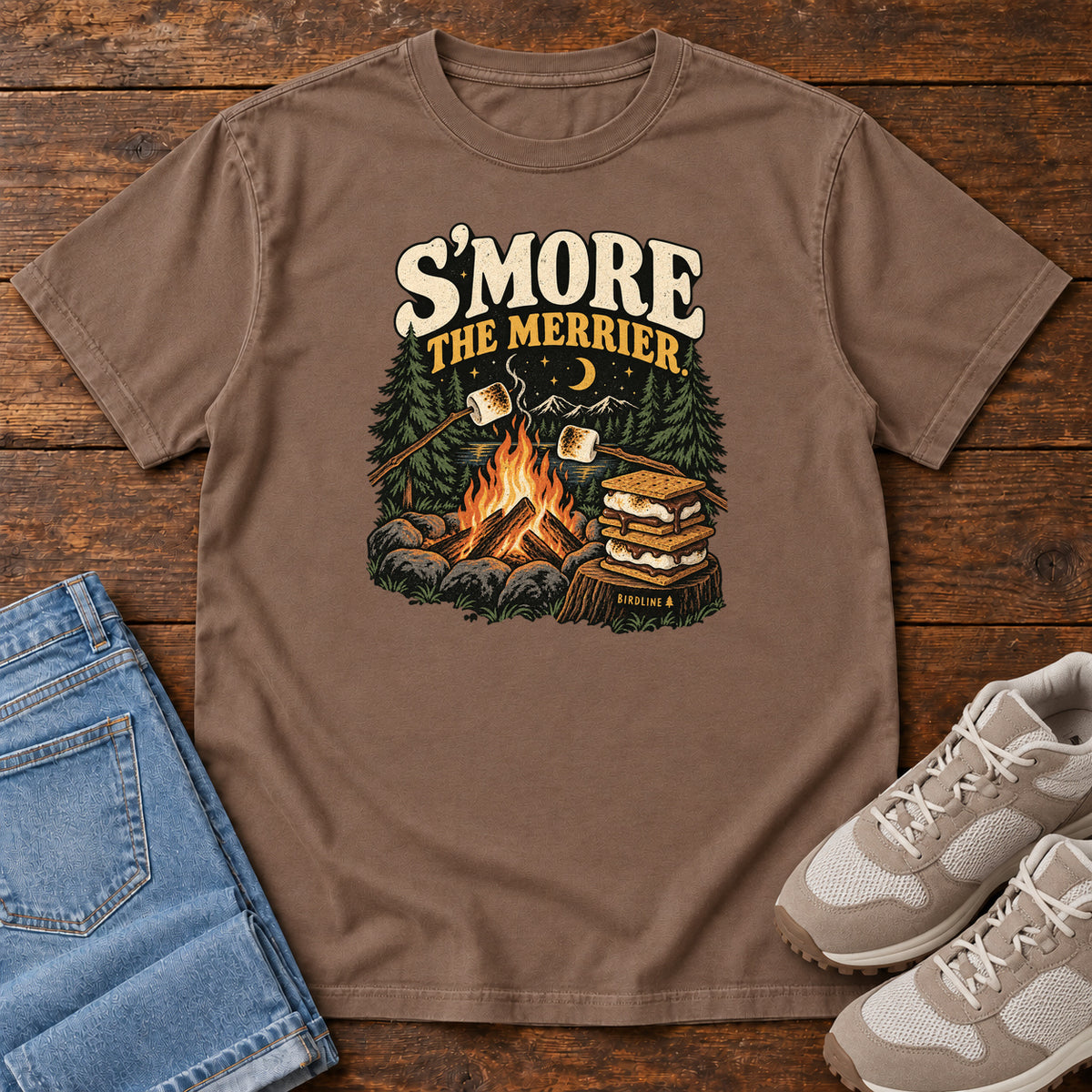 Espresso Smore the Merrier T-Shirt with a campfire, marshmallows on sticks and stacked s’mores beneath a crescent moon; front flat lay on wood.