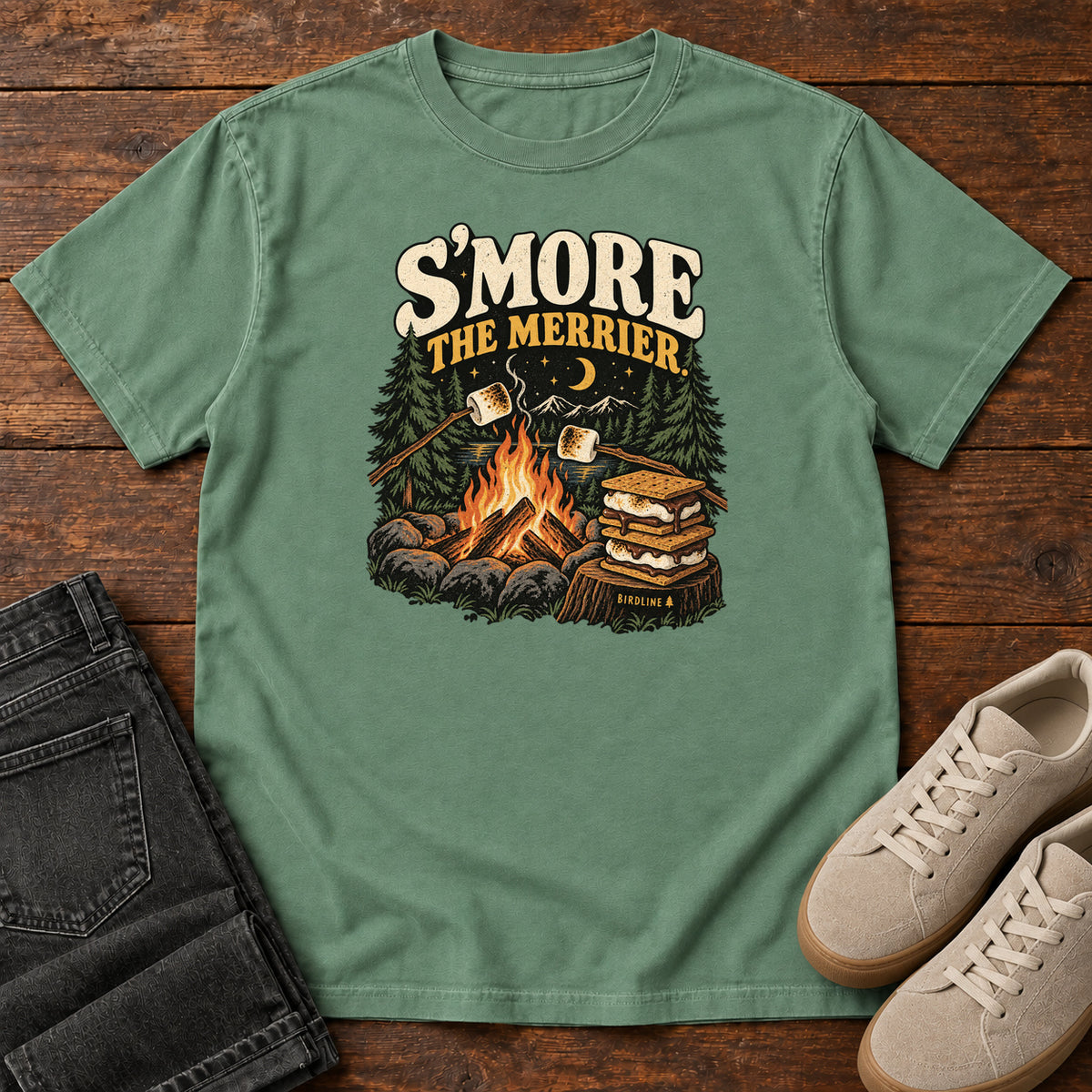 Light Green Smore the Merrier T-Shirt with a campfire, marshmallows on sticks and stacked s’mores beneath a crescent moon; front flat lay on wood.