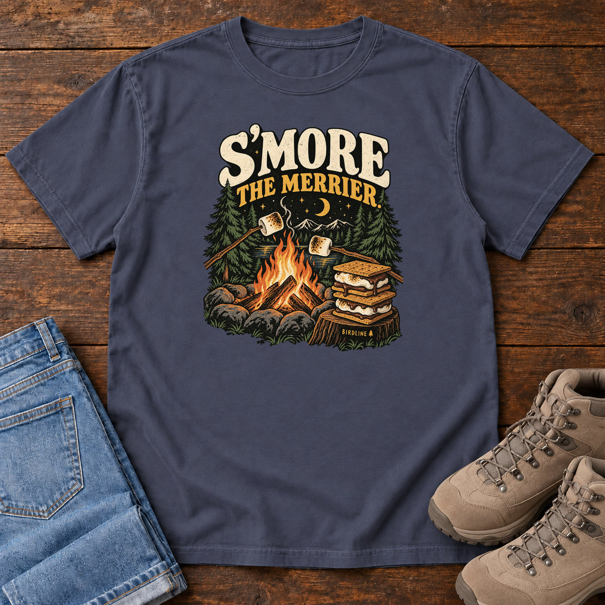 Navy Smore the Merrier T-Shirt with a campfire, marshmallows on sticks and stacked s’mores beneath a crescent moon; front flat lay on wood.