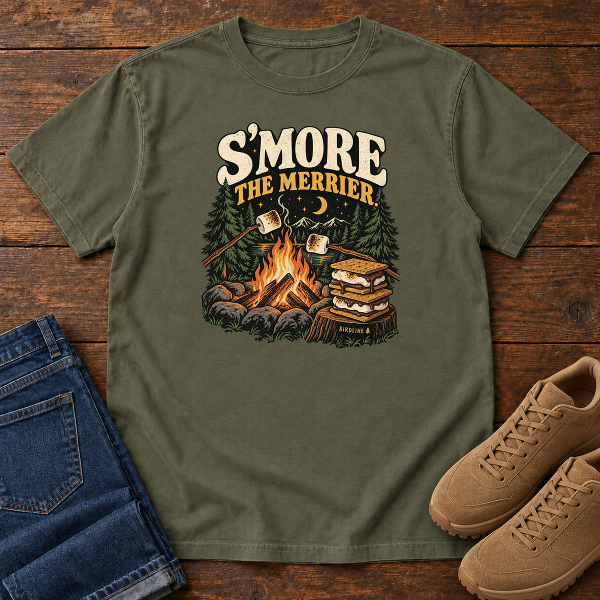 Sage Smore the Merrier T-Shirt with a campfire, marshmallows on sticks and stacked s’mores beneath a crescent moon; front flat lay on wood.