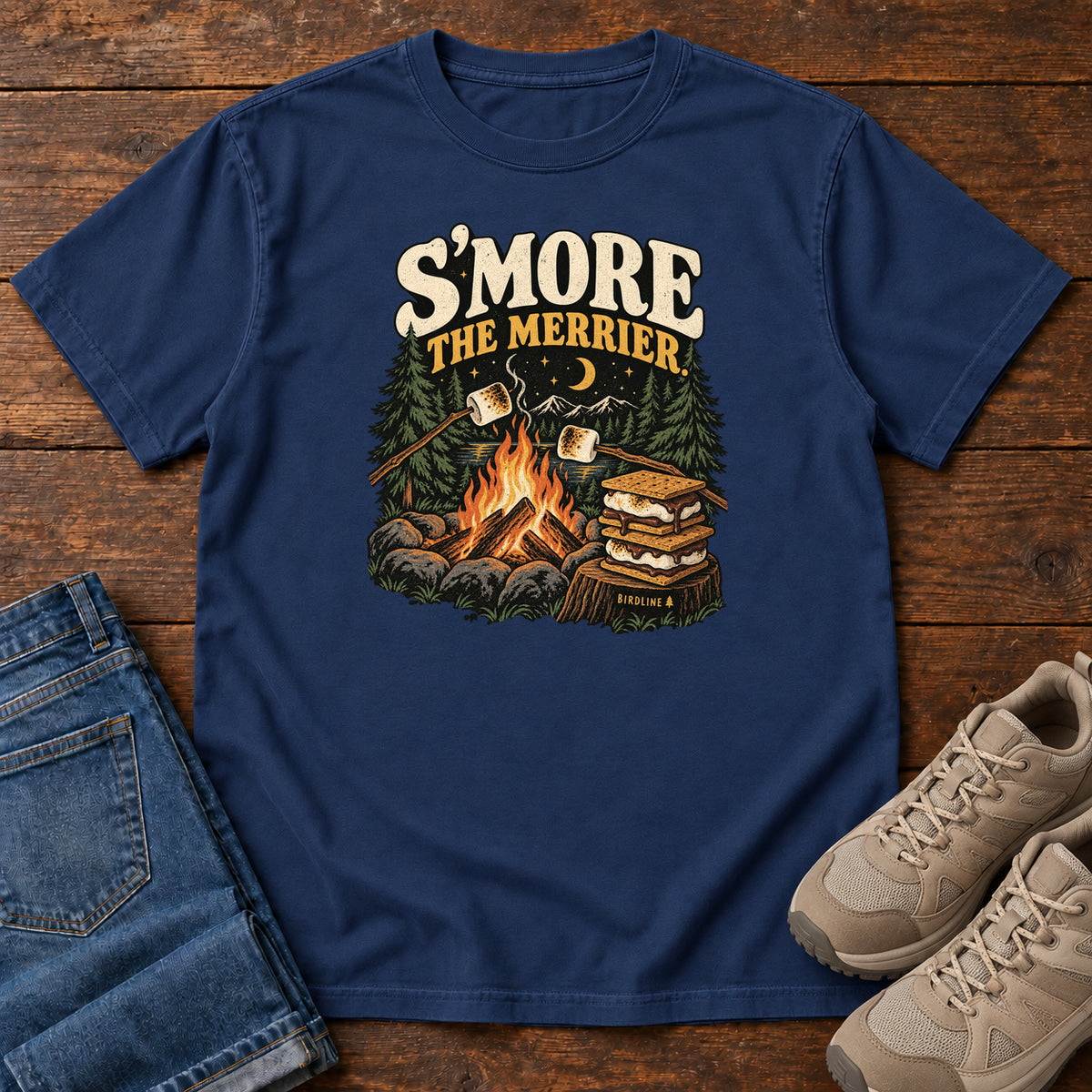 True Navy Smore the Merrier T-Shirt with a campfire, marshmallows on sticks and stacked s’mores beneath a crescent moon; front flat lay on wood.