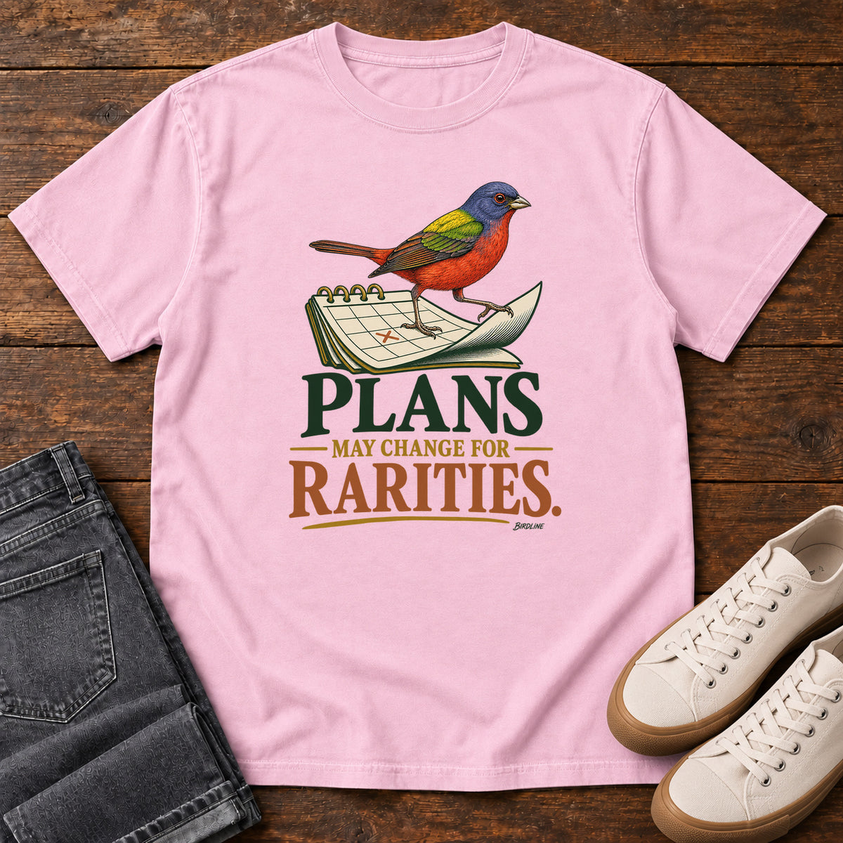 Blossom Plans May Change for Rarities T-Shirt with a red, blue and green bird perched on a curled calendar page; front flat lay on wood.