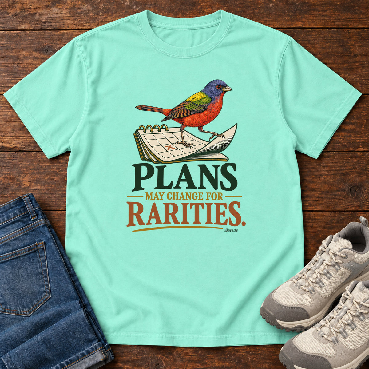 Chalky Mint Plans May Change for Rarities T-Shirt with a red, blue and green bird perched on a curled calendar page; front flat lay on wood.