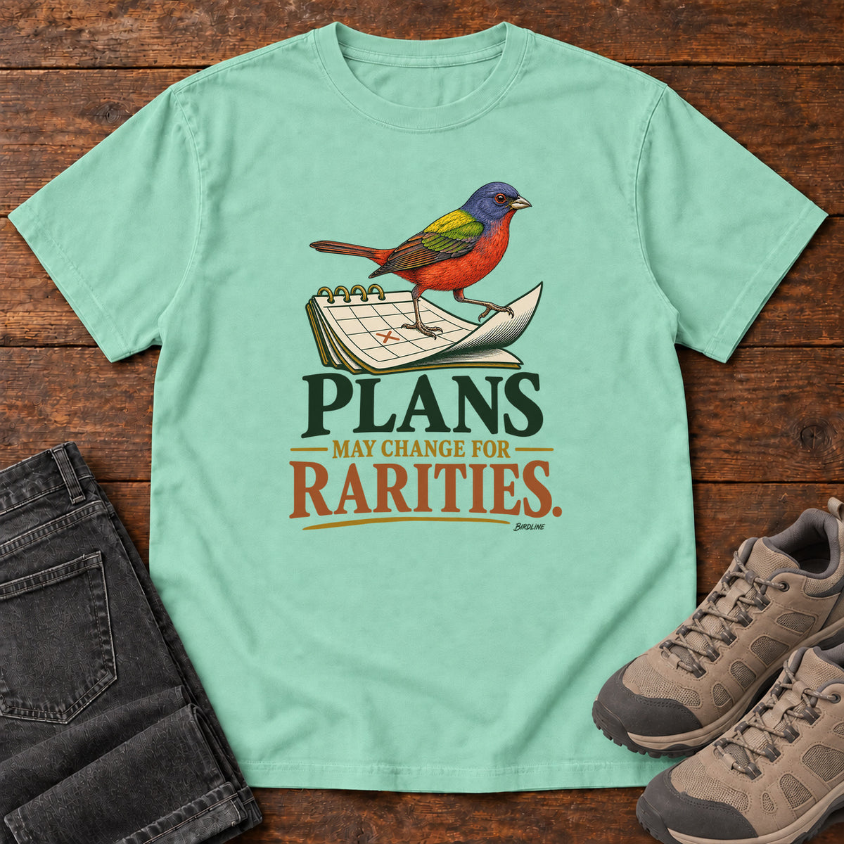 Island Reef Plans May Change for Rarities T-Shirt with a red, blue and green bird perched on a curled calendar page; front flat lay on wood.