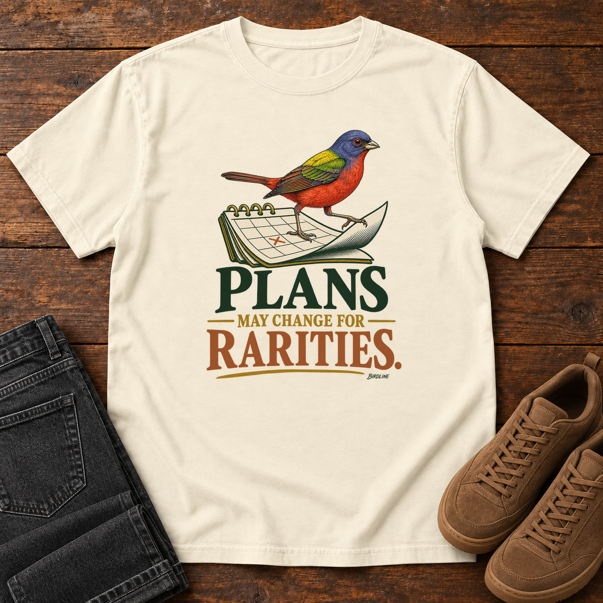 Ivory Plans May Change for Rarities T-Shirt with a red, blue and green bird perched on a curled calendar page; front flat lay on wood.