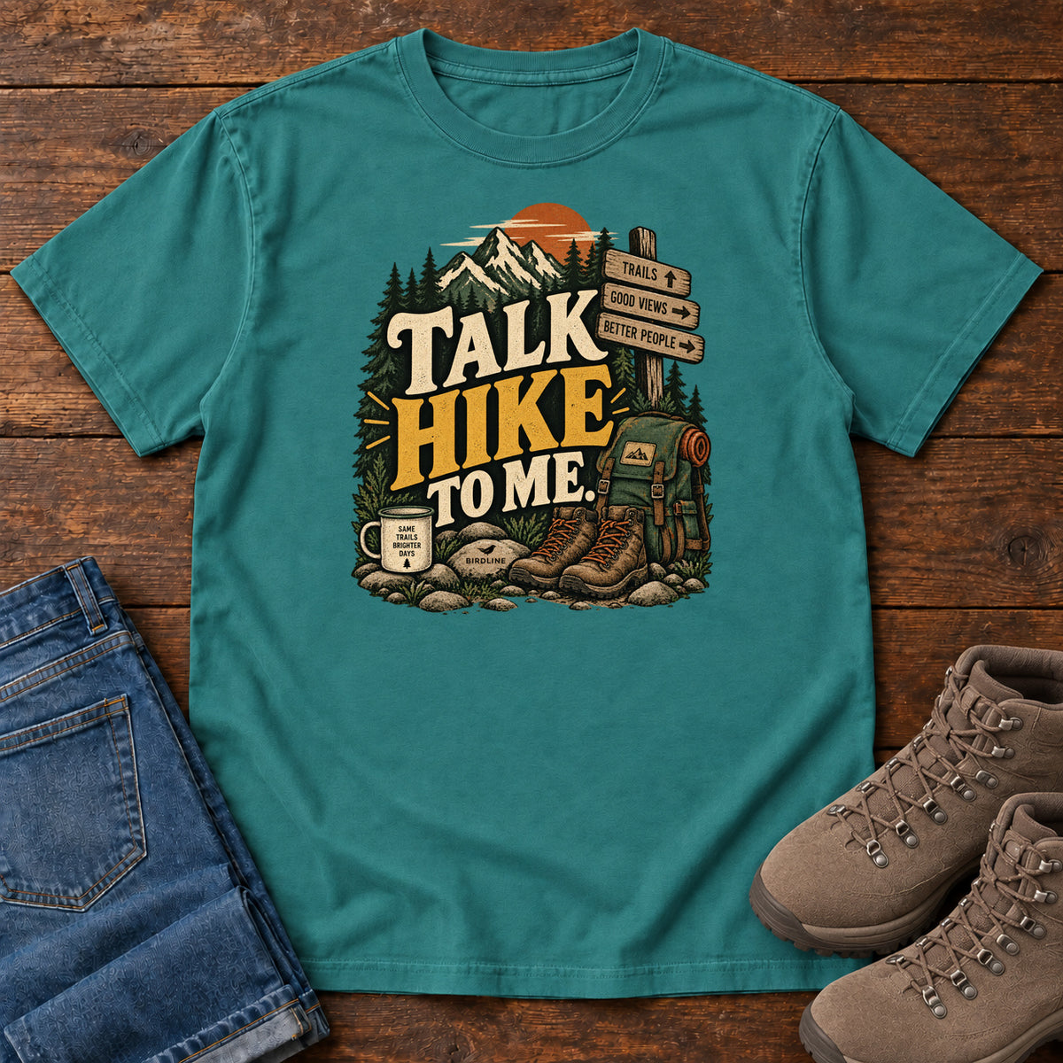 Emerald Talk Hike to Me T-Shirt with hiking boots, a backpack and trail sign beneath mountains and bold lettering; front flat lay on wood.