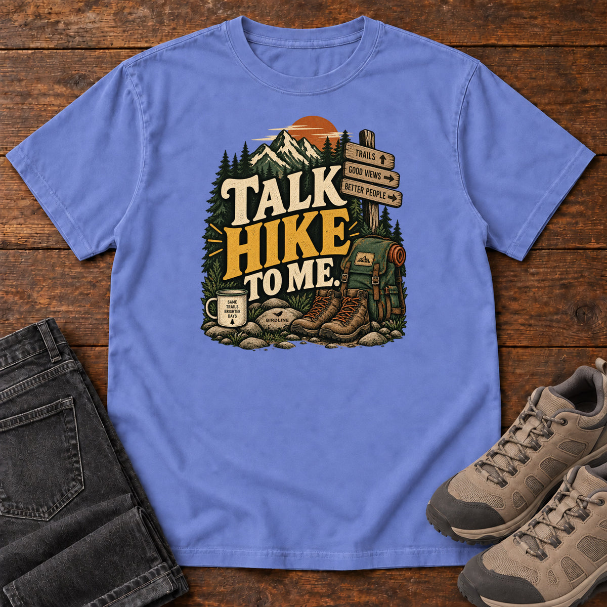 Flo Blue Talk Hike to Me T-Shirt with hiking boots, a backpack and trail sign beneath mountains and bold lettering; front flat lay on wood.