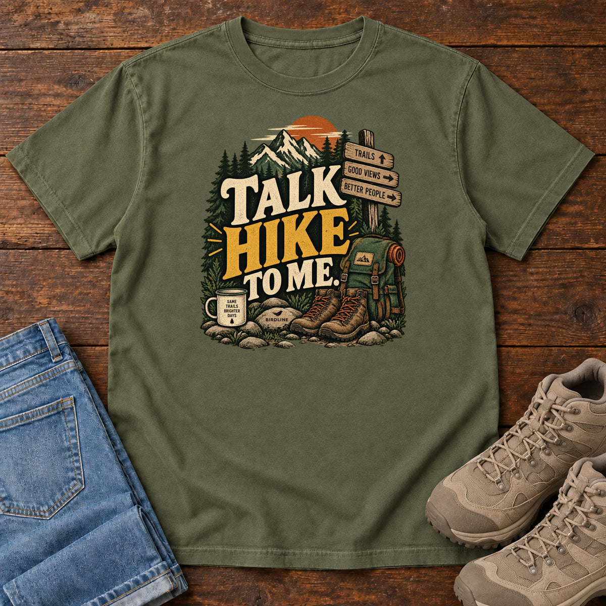 Hemp Talk Hike to Me T-Shirt with hiking boots, a backpack and trail sign beneath mountains and bold lettering; front flat lay on wood.