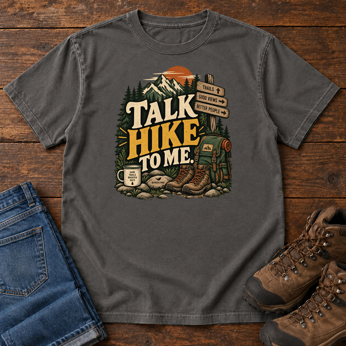 Pepper Talk Hike to Me T-Shirt with hiking boots, a backpack and trail sign beneath mountains and bold lettering; front flat lay on wood.