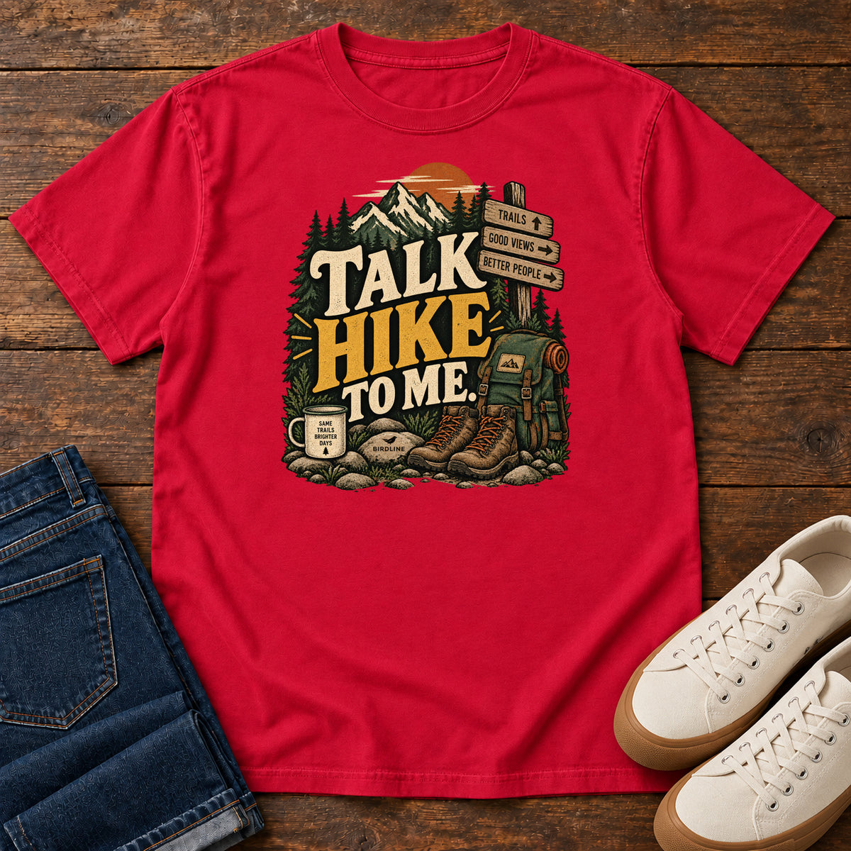 Red Talk Hike to Me T-Shirt with hiking boots, a backpack and trail sign beneath mountains and bold lettering; front flat lay on wood.
