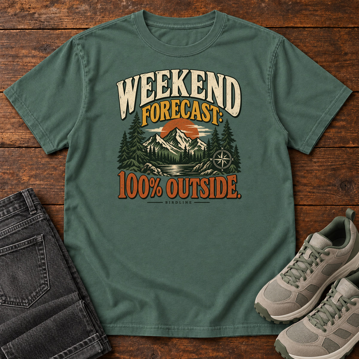 Blue Spruce Weekend Forecast 100 Percent Outside T-Shirt with mountains, evergreens, an orange sun and a compass beneath bold lettering; front flat lay on wood.